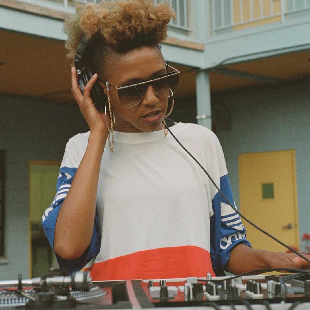 African-American woman DJ with sunglasses and headphones operating a sound mixer in an outdoor setting with natural lighting.