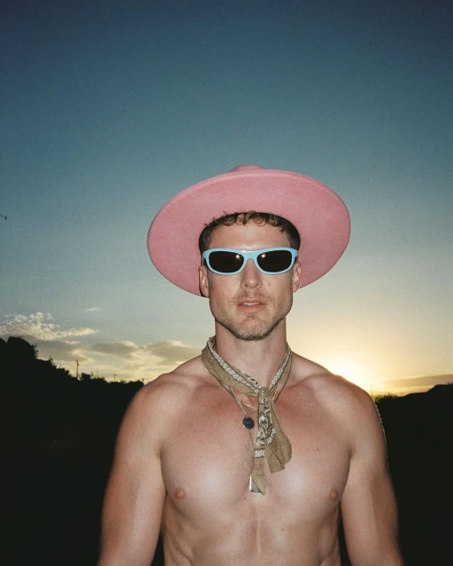 Portrait of a shirtless man wearing sunglasses and a pink hat against a sky with a setting sun. He also wears a patterned bandana.