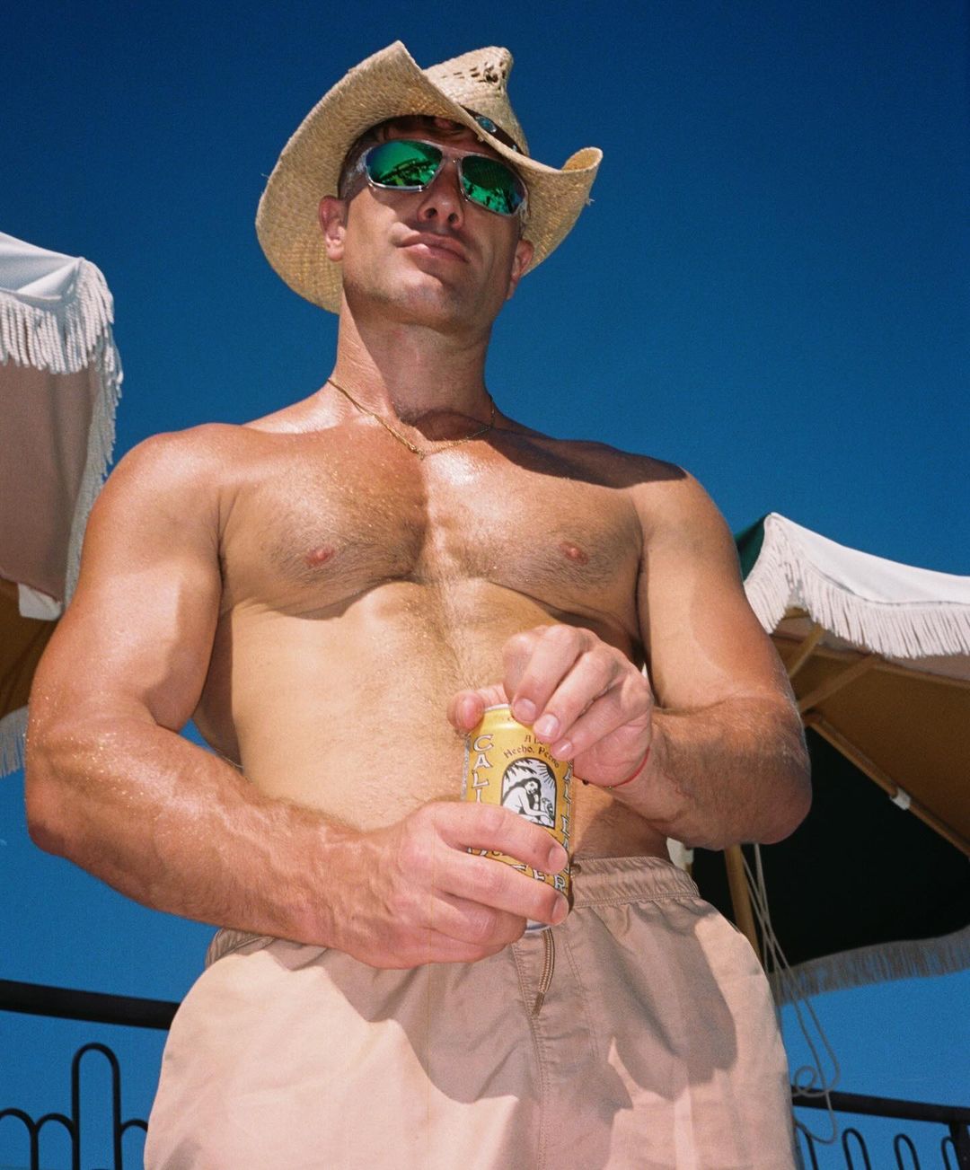 A man with bare torso, wearing a straw hat and sunglasses, is opening a beverage can under a clear blue sky with white umbrellas.