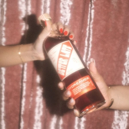 Two hands holding up a glass bottle of red beverage against a textured reddish background, captured by photographer Cully Wright.