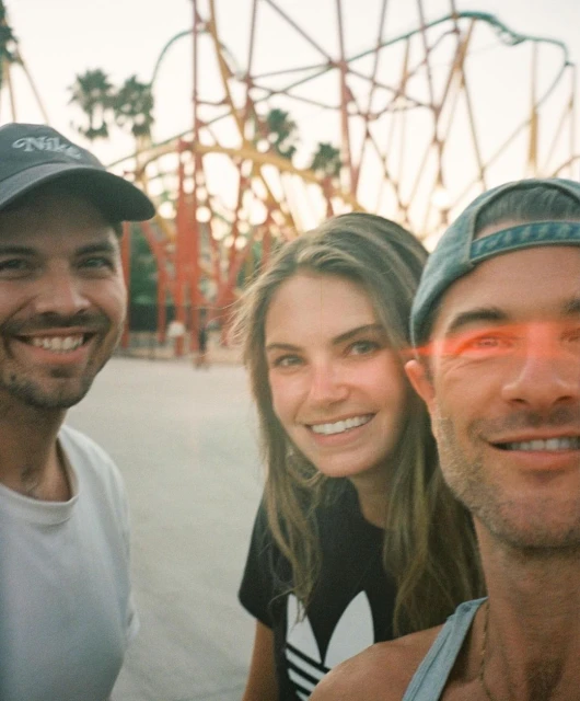 Three smiling people in a medium outdoor shot, wearing casual tops and hats, with a large rollercoaster in the background.