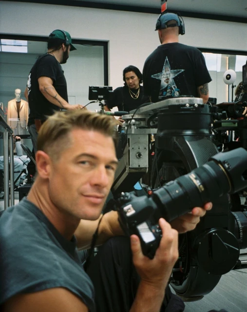 Man holds camera with film crew and equipment in the background. Indoor shoot features cameras, lighting, and a mannequin.