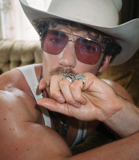 Man in cowboy hat and glasses, indoors. Rings on his fingers and a necklace around his neck. He's posing with a mustache.