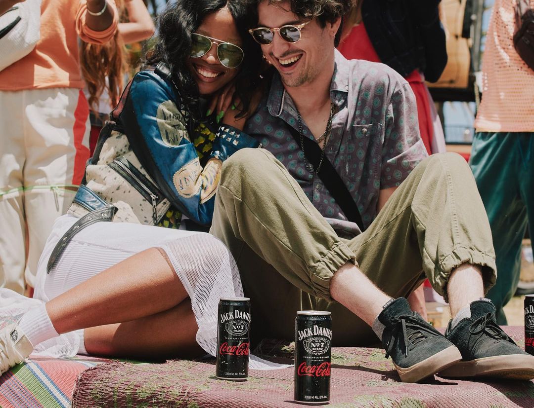 Two adults, an African American woman with sunglasses and a Caucasian man, sitting and smiling in outdoor setting with Jack Daniels and Coca-Cola.