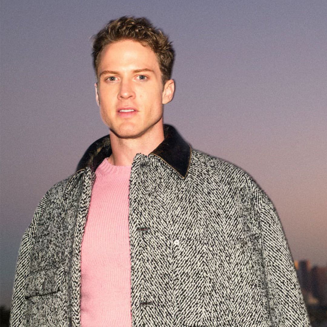 Portrait of a man wearing a black and white patterned jacket over a pink sweater against a sunset sky
