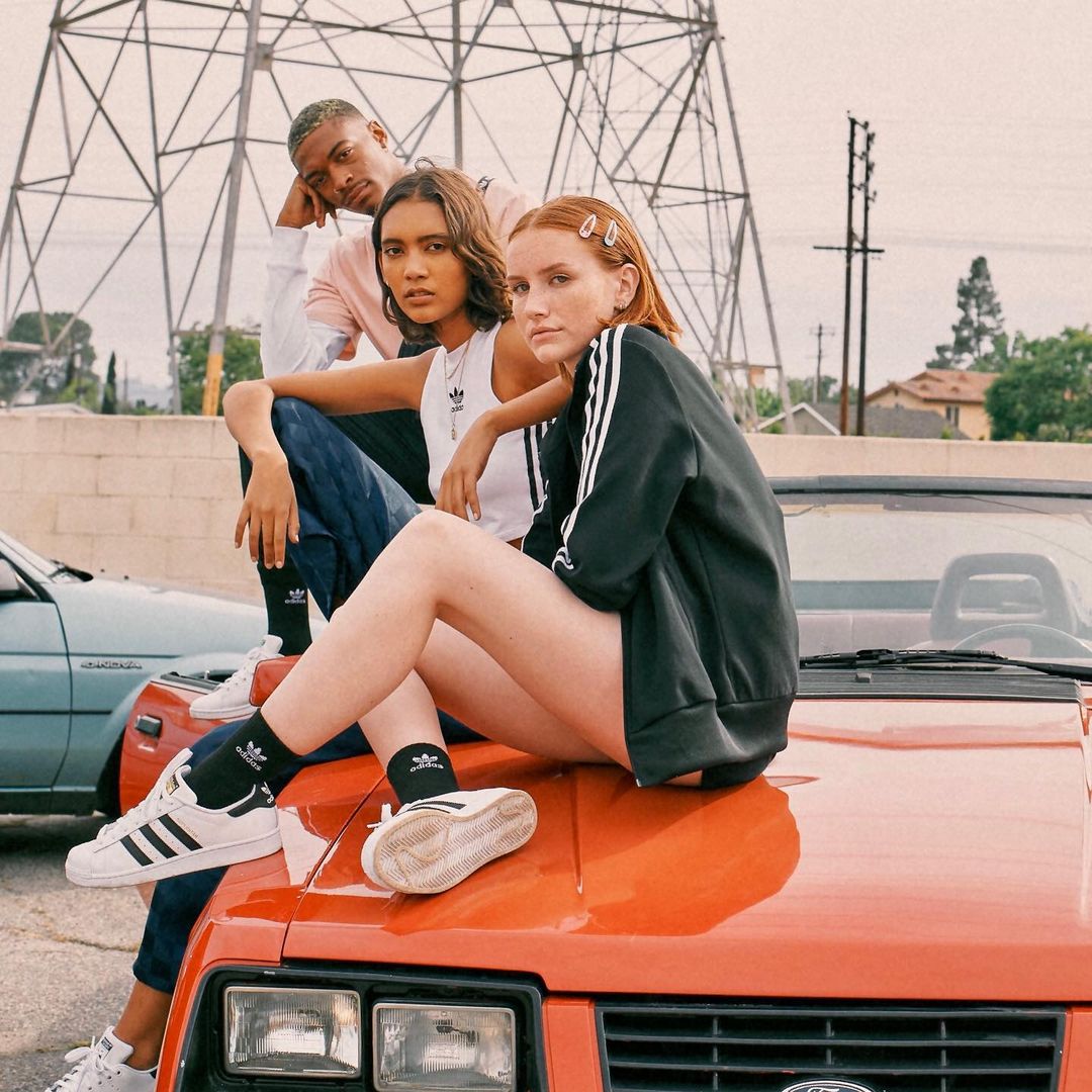 Three young adults in Adidas apparel pose on the hood of an orange car in front of power lines and a residential area on a cloudy day.