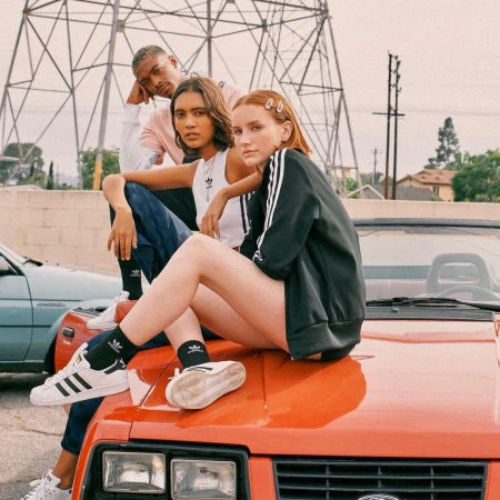 Three young adults in Adidas apparel pose on the hood of an orange car in front of power lines and a residential area on a cloudy day.