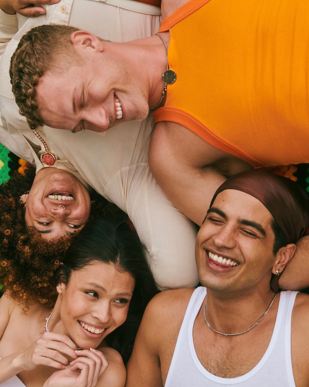A diverse group of four young adults smiles while lying head-to-head in a close-up, warm-toned shot highlighting their youthful faces.