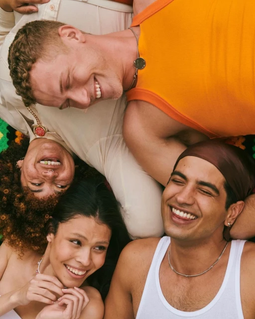 A diverse group of four young adults smiles while lying head-to-head in a close-up, warm-toned shot highlighting their youthful faces.