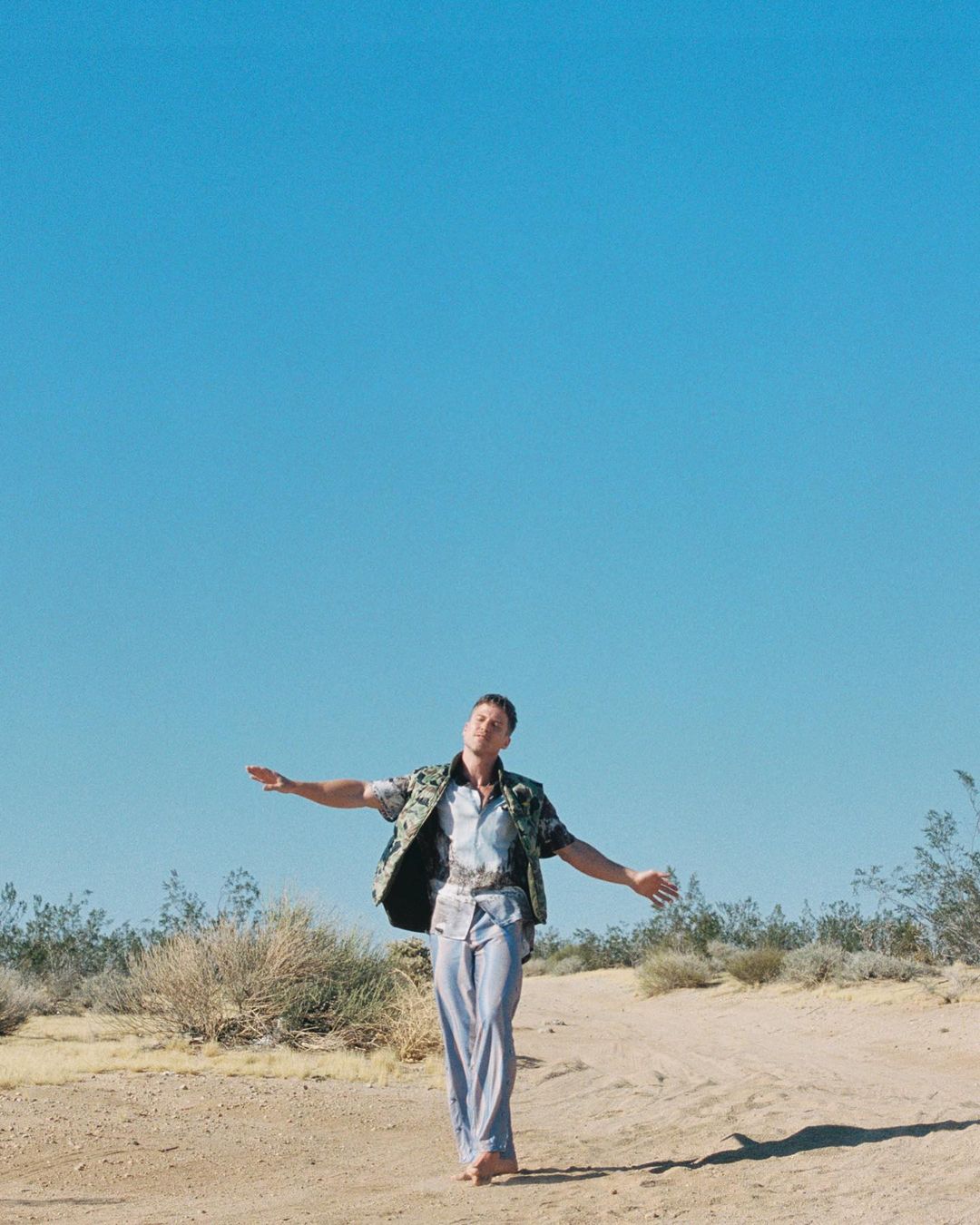 Adult male standing barefoot in desert terrain with arms outstretched under clear sky. Wearing casual shirt and pants with a vest.