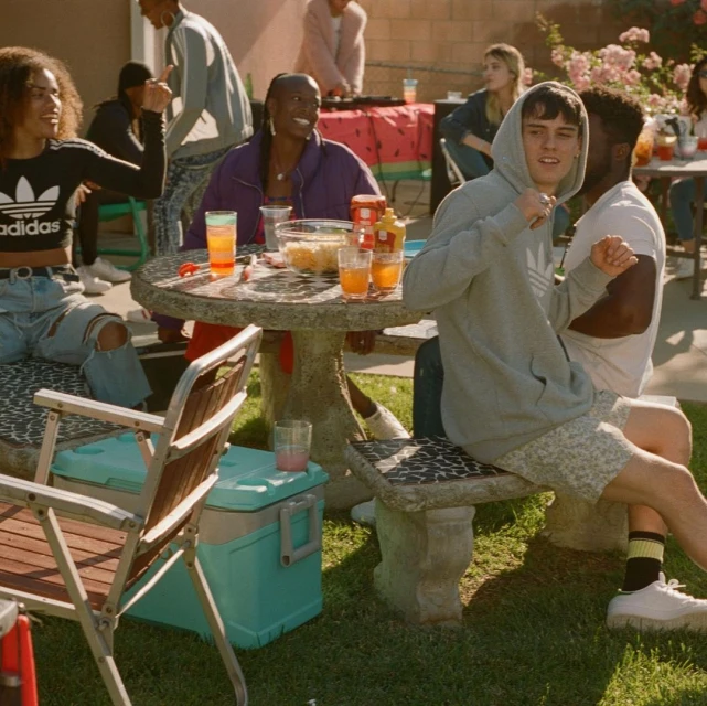 A group of young adults are gathered outside at a casual daytime social event, with tables of food and drinks, seated and standing in the yard.