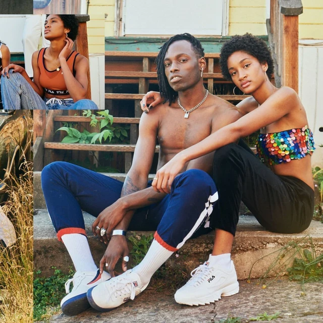 A couple poses on weathered wooden steps outside; the bare-chested man wears dark pants and white sneakers, his arm around the woman.