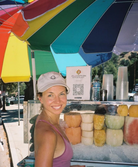 Woman smiling at camera next to a fruit stand with sliced fruit, underneath a colorful umbrella on a sunny day outdoors.