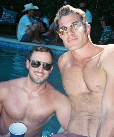 Two men pose shirtless by a pool with other adults visible in the background on a sunny day. Reflections in sunglasses.