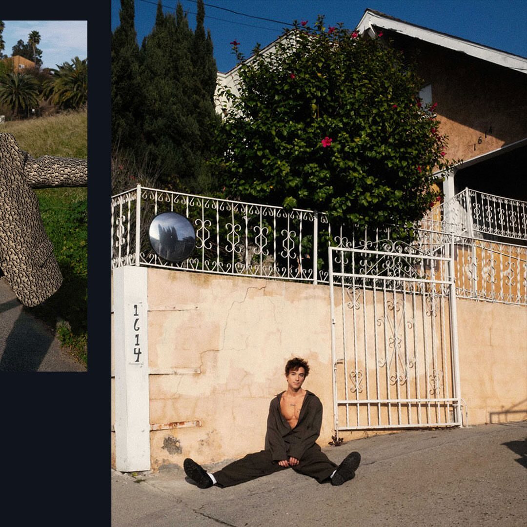 Man sits in front of wall and gate with house in background. Dark green jumpsuit, with chest exposed, black shoes, and short dark hair.