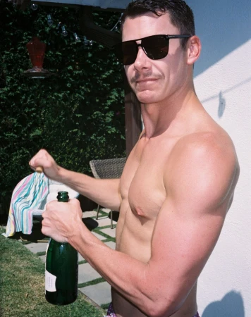 Shirtless man wearing sunglasses holding a green champagne bottle outdoors, photographed by Cully Wright.