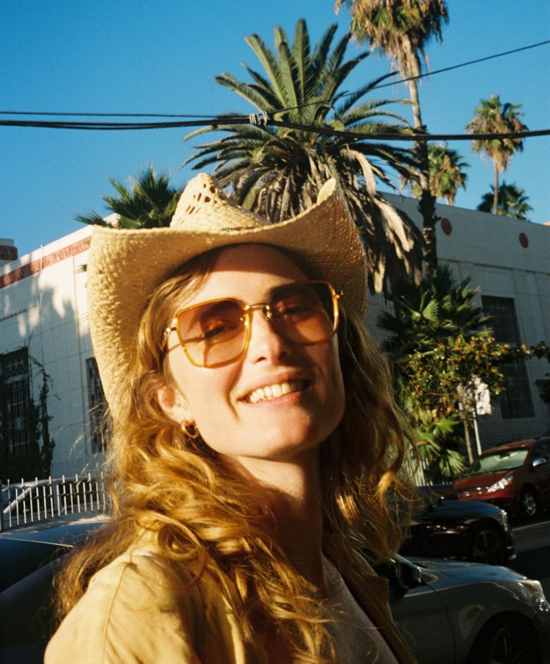 Smiling woman in cowboy hat and sunglasses stands outside on city street with palm trees and building in background on a sunny day.