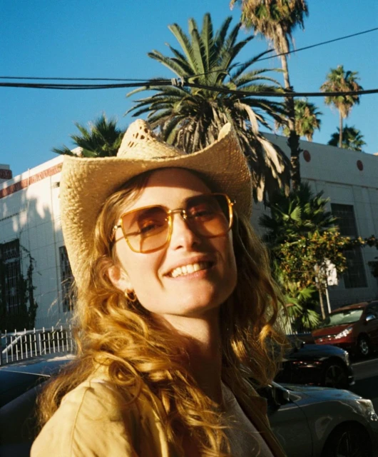 Smiling woman in cowboy hat and sunglasses stands outside on city street with palm trees and building in background on a sunny day.