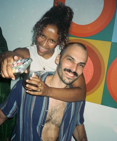 Smiling woman in white tank top embracing a man in a striped shirt holding a drink in New York by Cully Wright.
