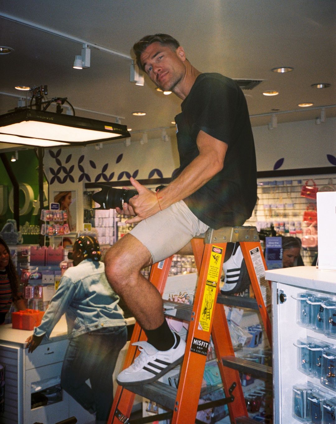 Man with camera sits atop a ladder in a retail environment with fluorescent lighting. Two women shop in the background.