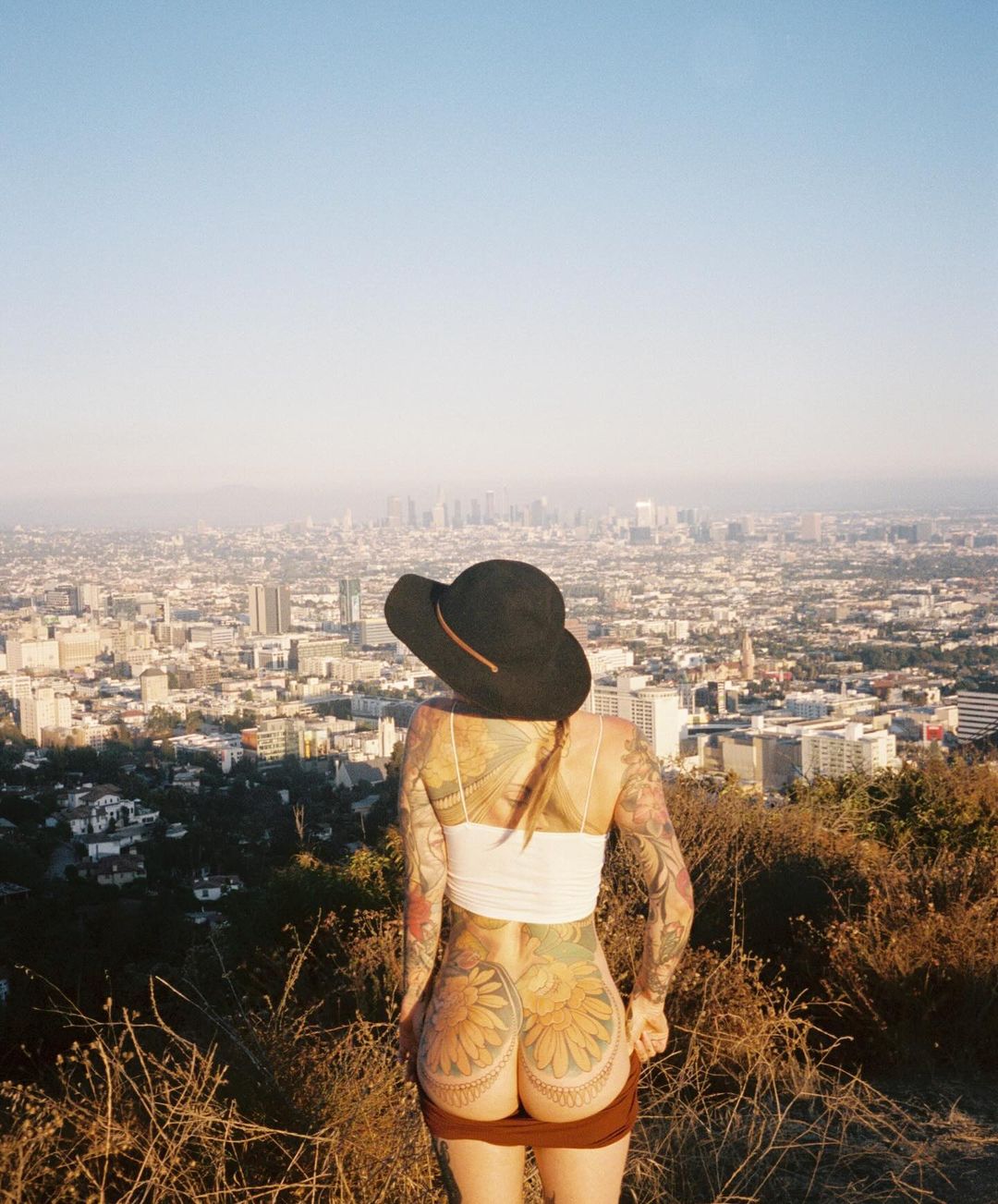 Tattooed woman in hat stands atop hill overlooking city skyline. Woman wears skirt and white top, with tattoos on her back and arms.