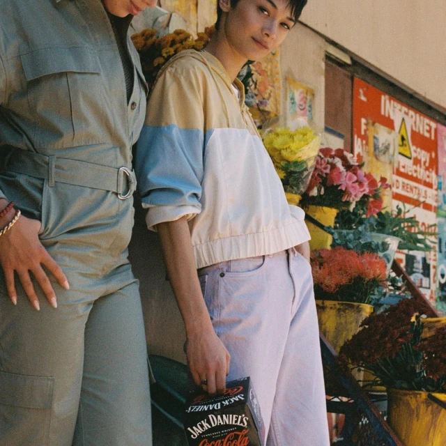 Two young women stand outdoors in casual outfits next to flowers. One holds a Jack Daniel's & Coca-Cola can in a medium shot.