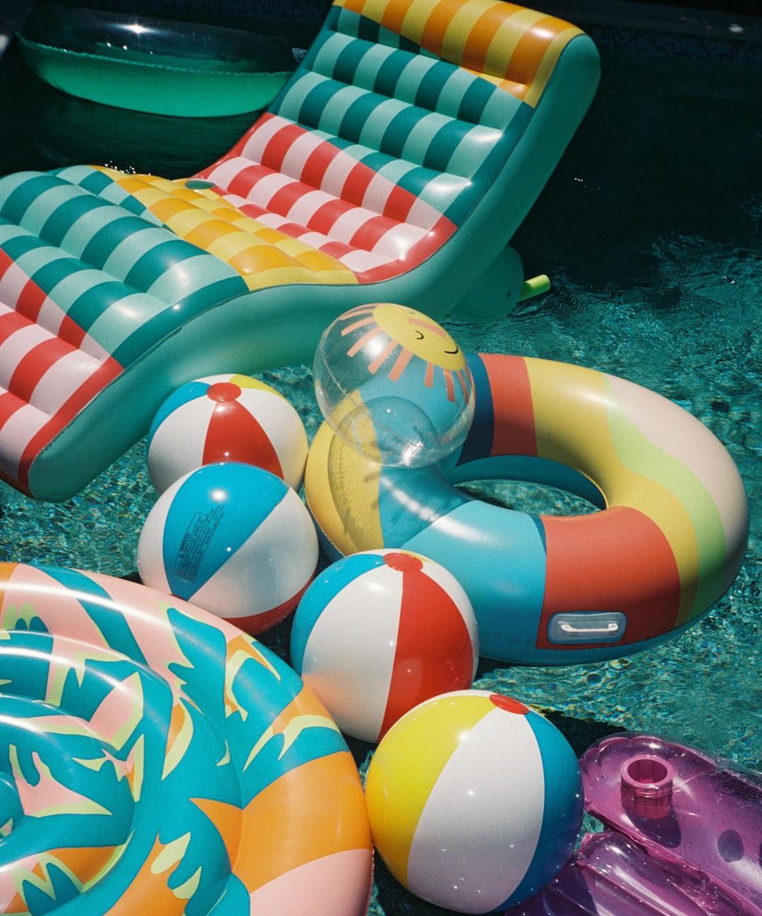 A collection of colorful inflatable pool toys including balls, rings, and a floating lounge chair in a swimming pool setting.