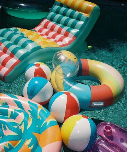 A collection of colorful inflatable pool toys including balls, rings, and a floating lounge chair in a swimming pool setting.