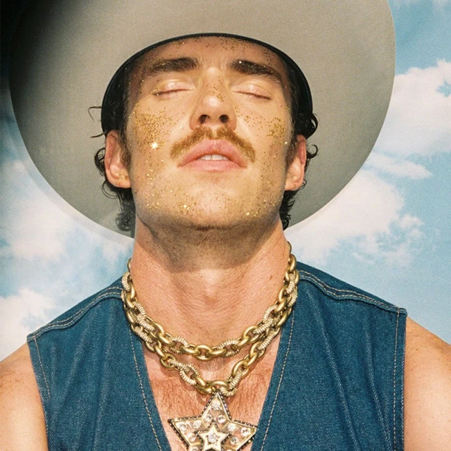 Close-up of a man with eyes closed, wearing a cowboy hat and gold chains, against a blue sky with clouds backdrop. Gold glitter on face.