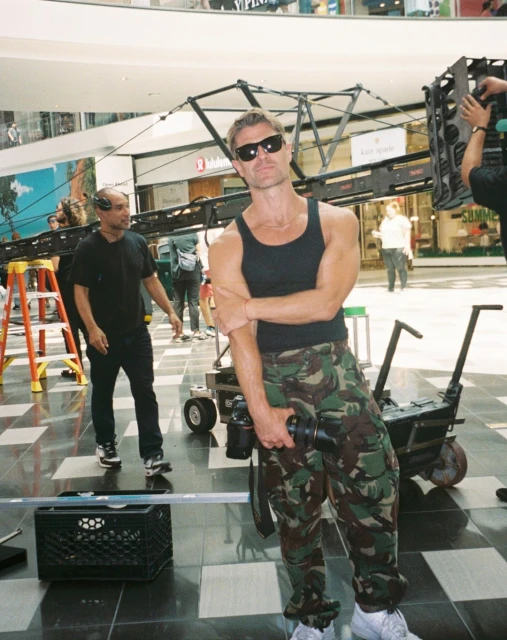 Man in tank top and camouflage pants poses with camera equipment indoors, with another man and retail shops in the background.