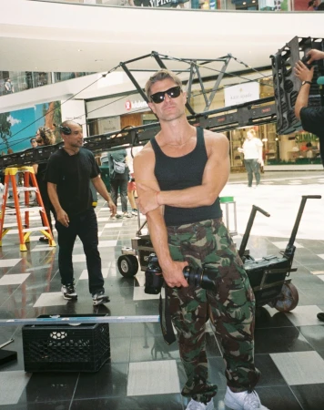 Man in tank top and camouflage pants poses with camera equipment indoors, with another man and retail shops in the background.