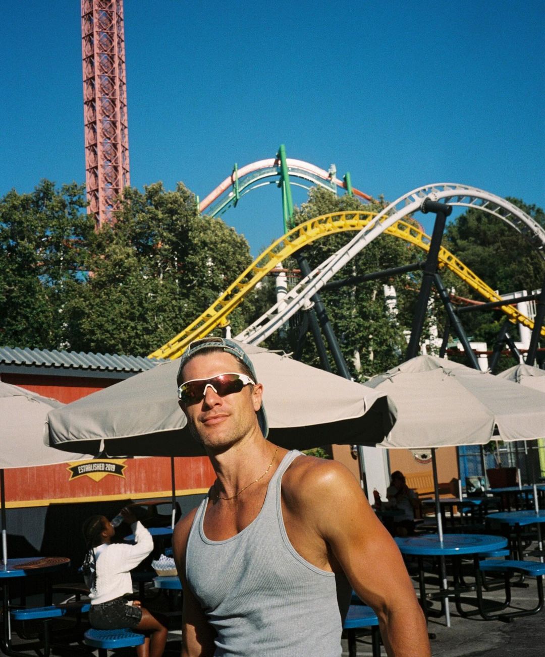 Man smiles in a gray tank top with sunglasses, at an amusement park with roller coasters and a tall tower ride in the background.