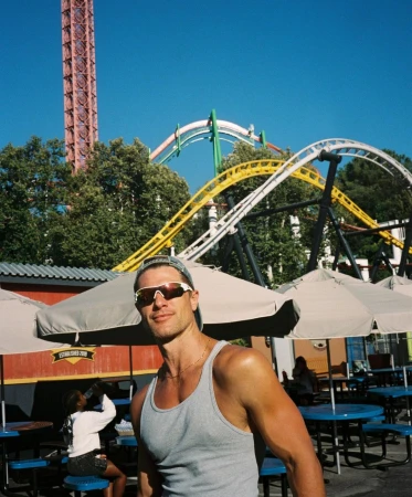 Man smiles in a gray tank top with sunglasses, at an amusement park with roller coasters and a tall tower ride in the background.
