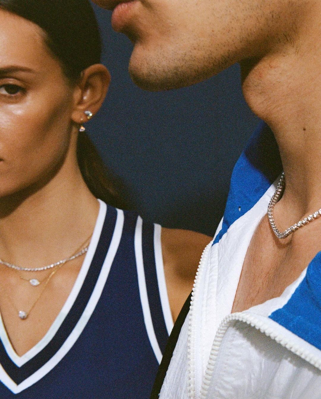 Close-up of a stylish couple with diamond jewelry, one wearing a navy tank top and the other in a white and blue athletic jacket.