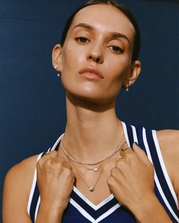 Close-up portrait of a woman wearing layered necklaces and a blue-and-white striped tank top against a dark blue background.