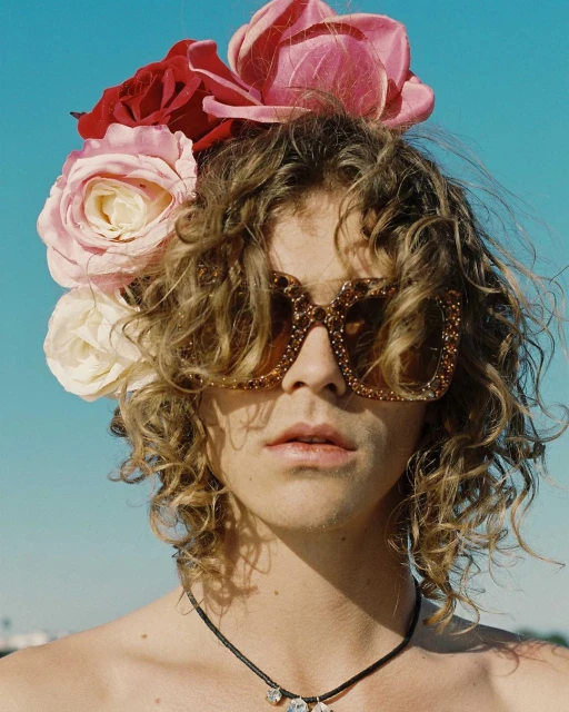 Person with curly hair wearing large sunglasses and floral headpiece against a bright blue sky in an outdoor setting.