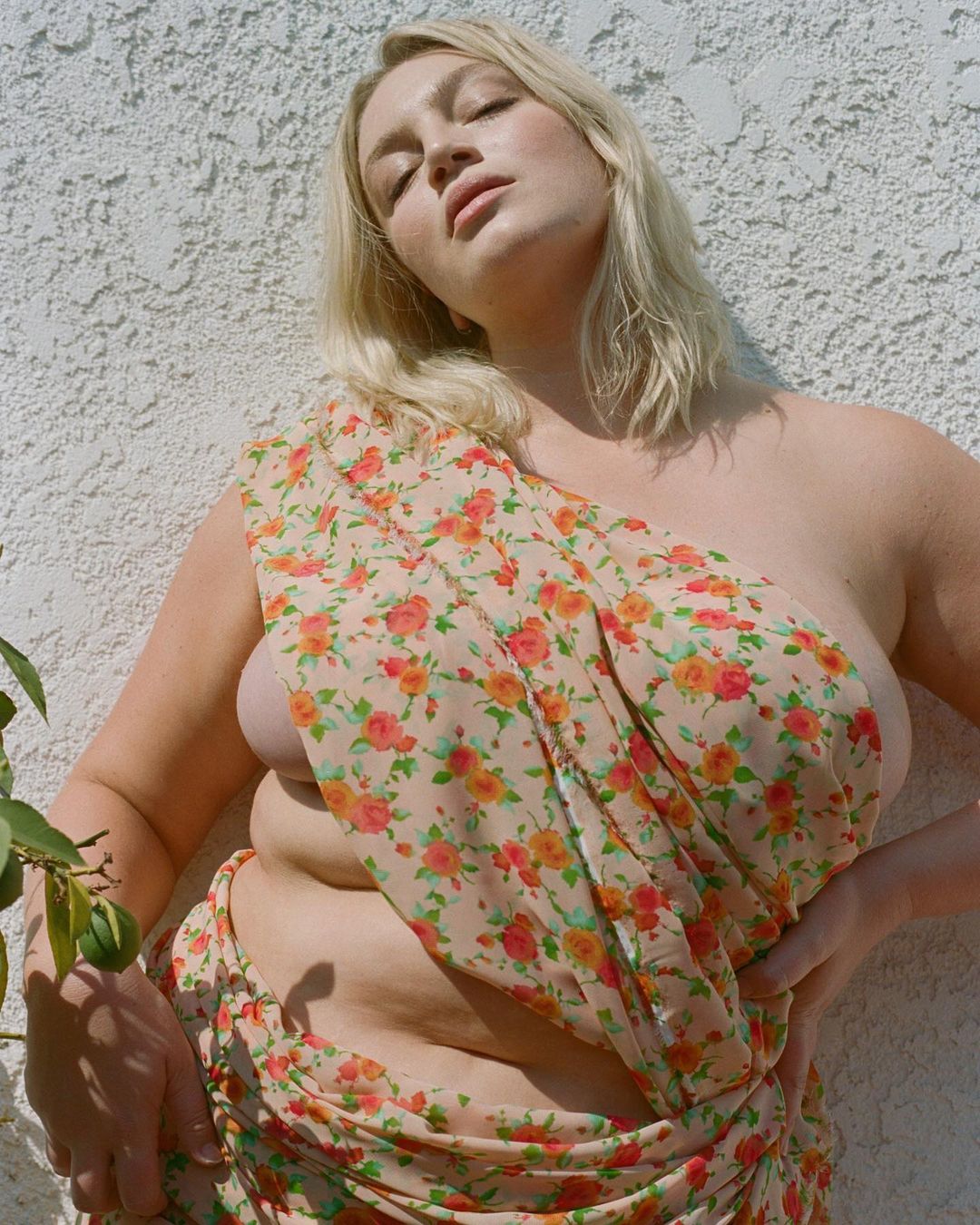 Plus size blonde woman wearing a floral sheer wrap leaning against a textured white stucco wall outdoors in bright sunlight.