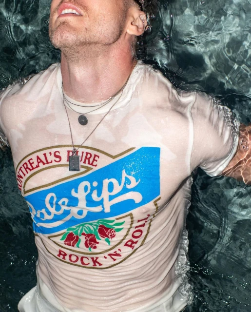 Man with wet white t-shirt floats in water. The shirt has a vintage-style graphic with the text 'Montreal's Pure Babe Lips, Rock 'n' Roll'.