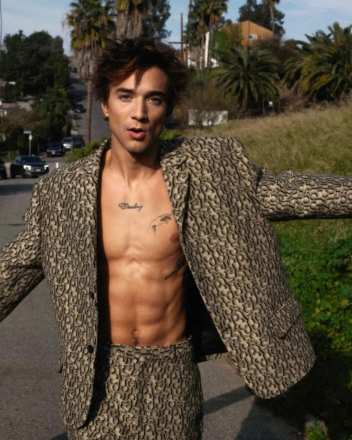 A young adult man in a patterned suit poses outdoors with arms outstretched, showing his torso, with a road and greenery in the background.