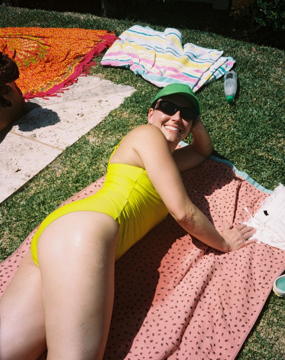 A woman in a yellow swimsuit lies on a patterned towel on grass wearing a green hat and sunglasses smiling with other towels visible.