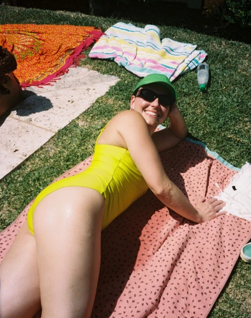 A woman in a yellow swimsuit lies on a patterned towel on grass wearing a green hat and sunglasses smiling with other towels visible.