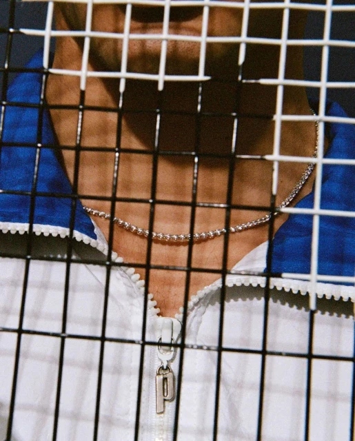 Close-up of a person wearing a silver chain necklace and a white jacket with blue accents, seen through a metal fence.