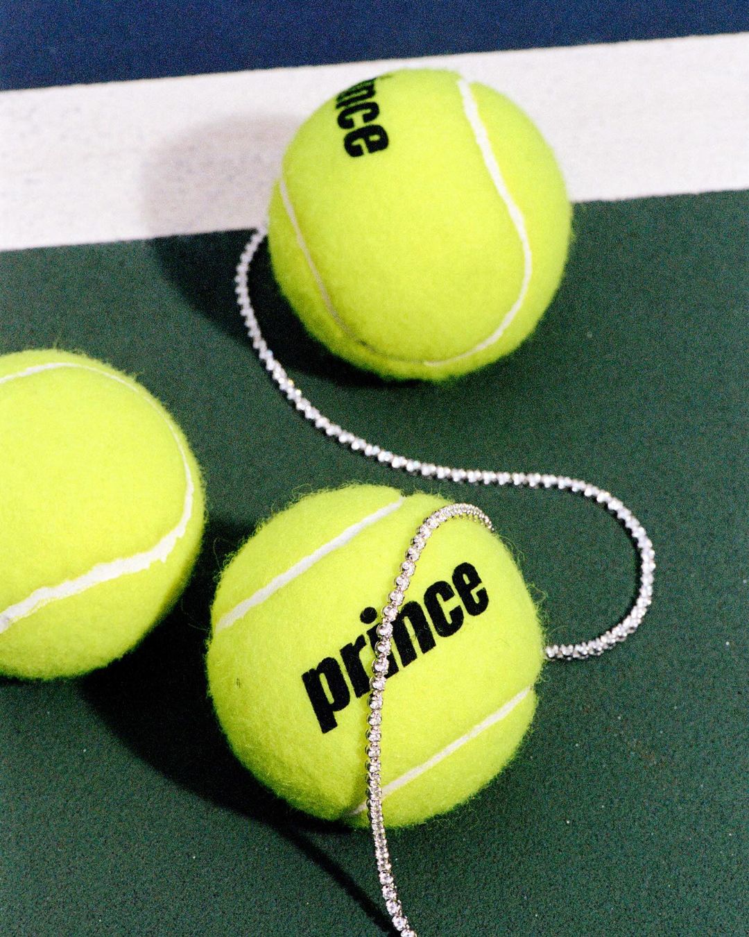 Close-up of three tennis balls on a green court with white lines, one wrapped with a diamond tennis bracelet and the Prince logo.