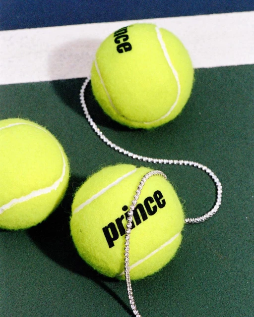 Close-up of three tennis balls on a green court with white lines, one wrapped with a diamond tennis bracelet and the Prince logo.