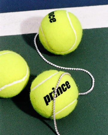 Close-up of three tennis balls on a green court with white lines, one wrapped with a diamond tennis bracelet and the Prince logo.