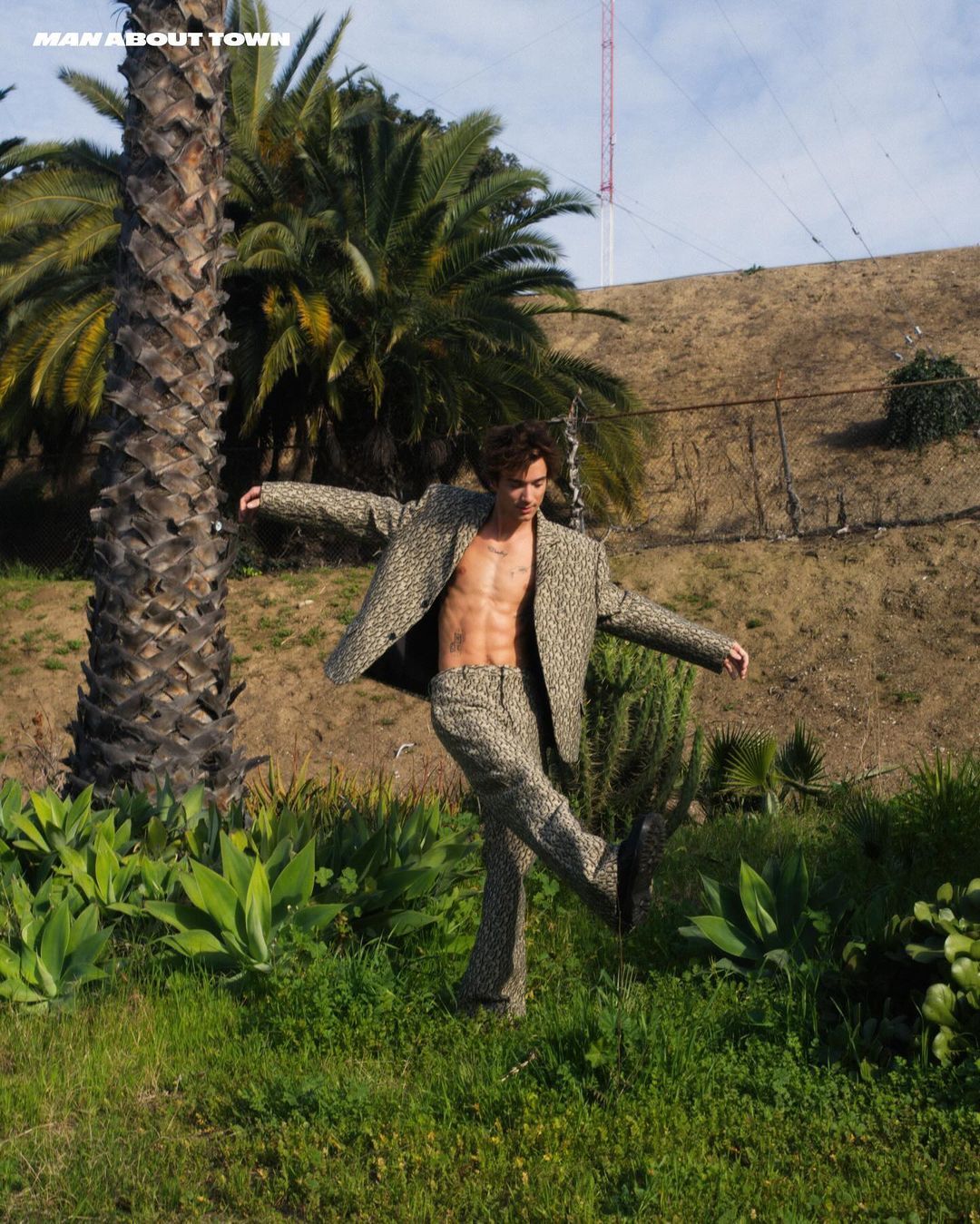 Man poses outdoors wearing tan patterned suit, with palm trees and hillside in background. Fashion editorial style shot.