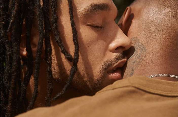 Close-up of two adult males embracing outdoors, one with dreadlocks and eyes closed, the other in a tan shirt, showcasing intimacy.