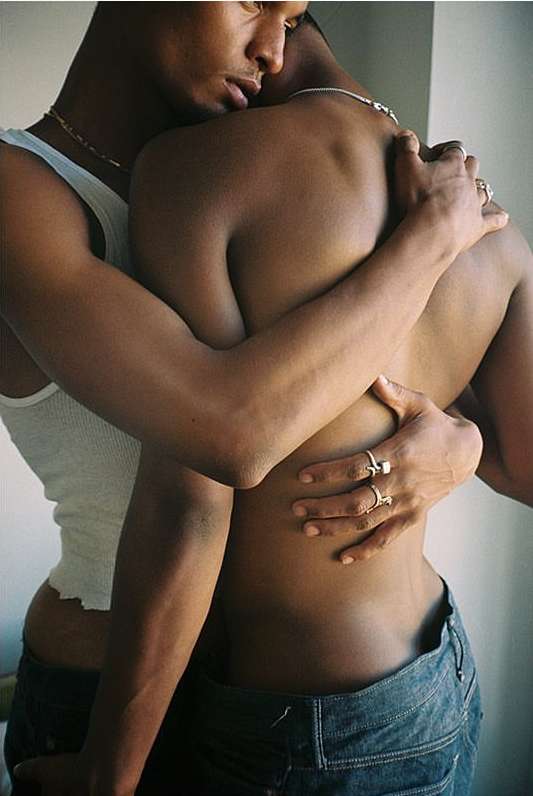Close-up of two dark-skinned adult males embracing indoors; one wears a white tank top and blue jeans, the other has bare torso and jeans.