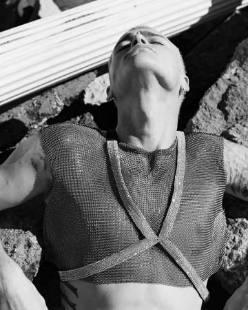 Monochrome portrait of a person with short blonde hair wearing a chainmail top, head tilted back against rocky rubble.