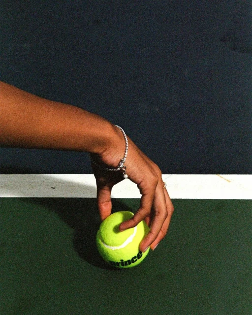 Close-up shot on a tennis court featuring a light yellow tennis ball being held by a female hand wearing a bracelet and a ring.